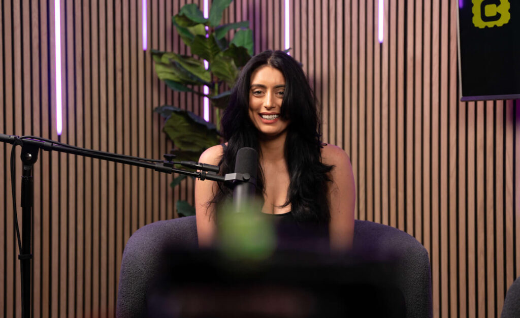 The Content Factory A person with long dark hair sits in a podcast studio in Austin, Texas, with a microphone in front of them, smiling. The background features vertical wooden slats and pink neon lights. In Austin, Texas